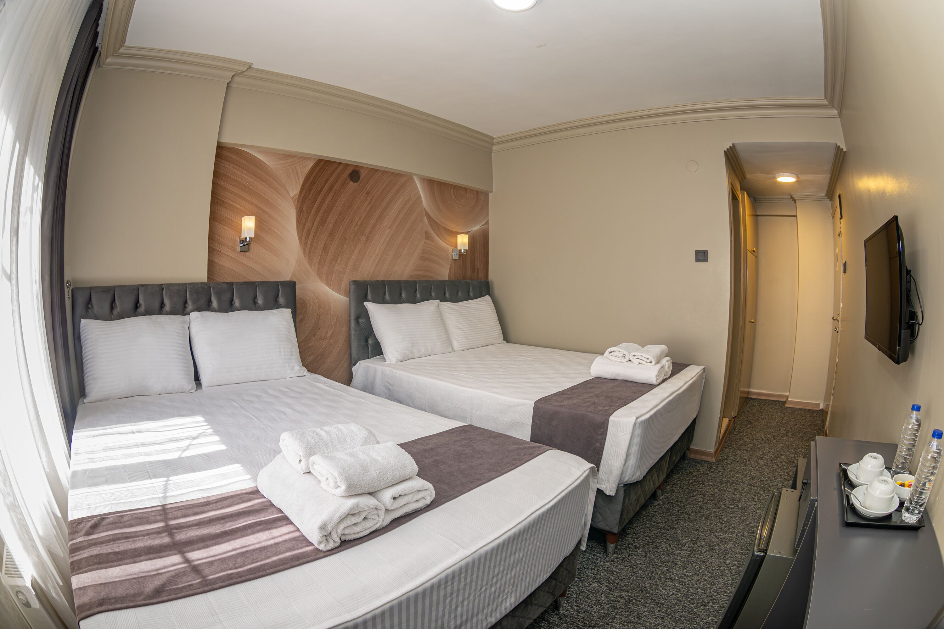 A hotel room with two beds, a flat-screen TV, a desk with a tray of toiletries, and a wooden accent wall.
