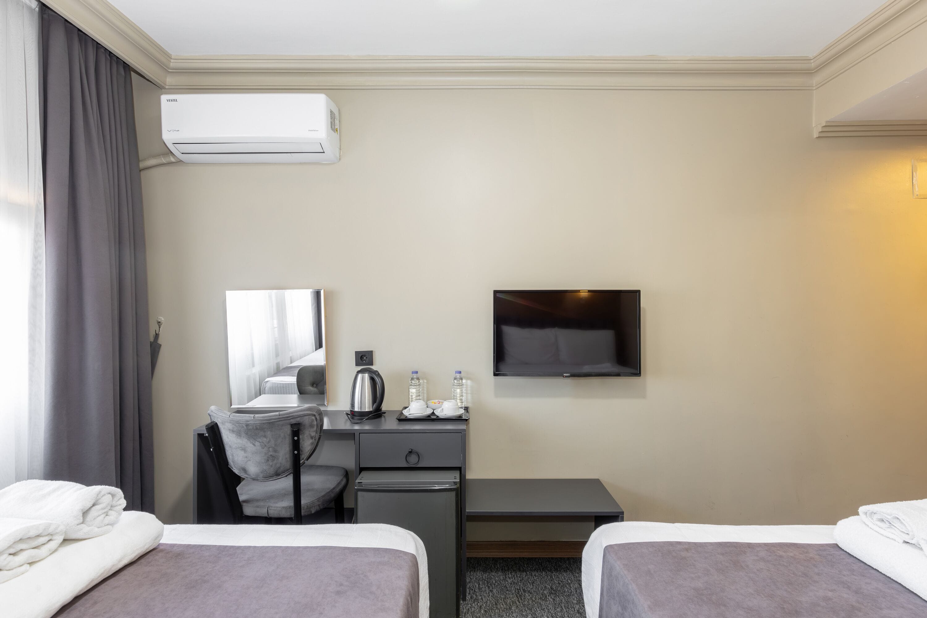 A hotel room with two beds, a desk, a chair, a flat-screen TV, an air conditioner, and a window with curtains.