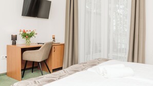 A hotel room with a bed, a desk with a chair, a TV mounted on the wall, and a window with curtains. - Hotel Anna Villa (Balatonfoldvar)