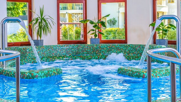 An indoor swimming pool with mosaic tile edges, potted plants, and a view of buildings through large windows. - Hotel Anna Villa (Balatonfoldvar)