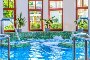 Una piscina interior con bordes de mosaicos, plantas en macetas y vistas a edificios a través de amplias ventanas.