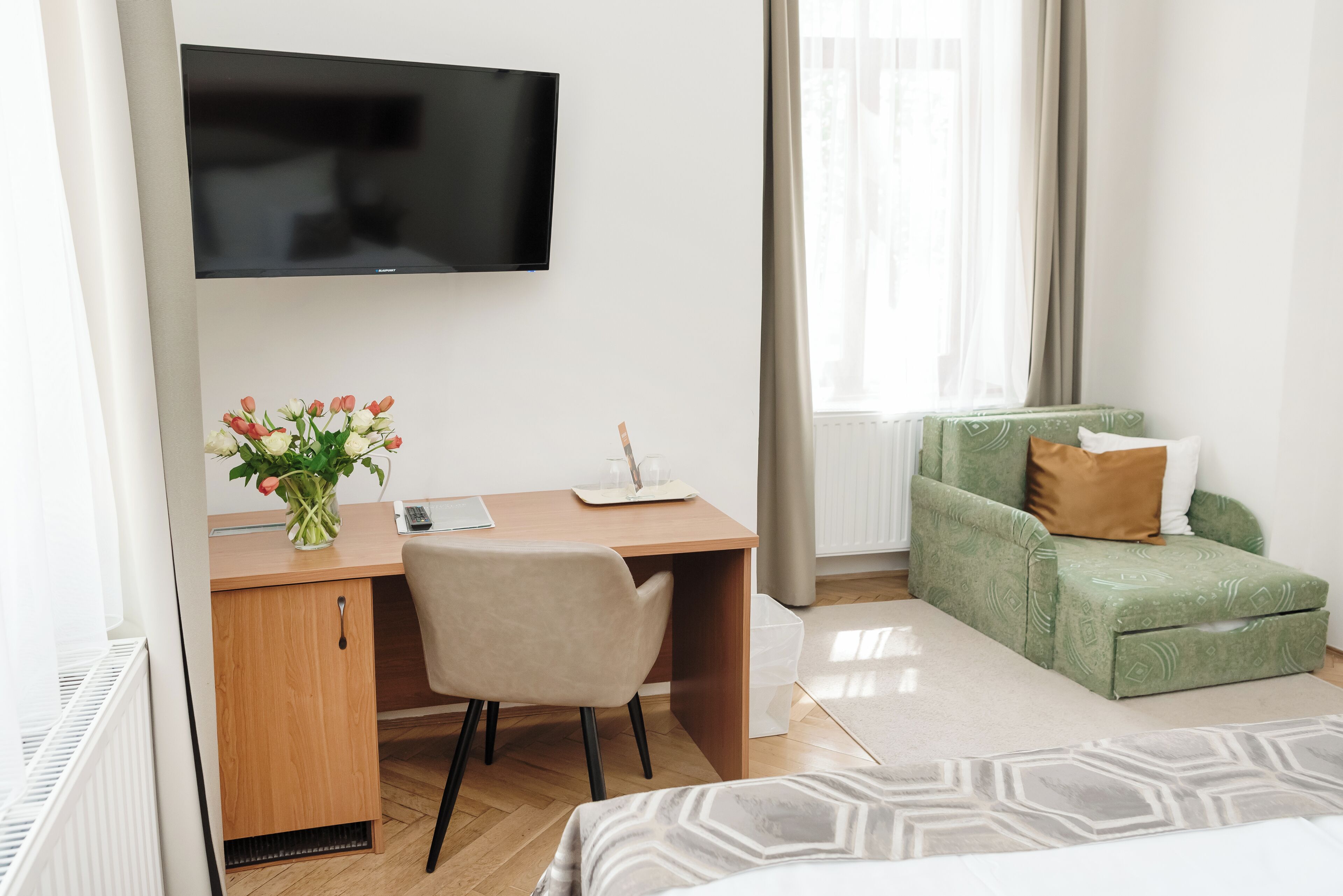 Quarto de hotel com escrivaninha de madeira, uma cadeira, cama com colcha estampada, sofá verde com almofadas, TV de tela plana na parede e janela com cortinas leves.