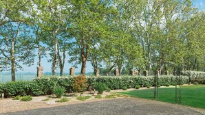 A paved garden area with neatly trimmed hedges and trees, a clear sky, and a distant view of the sea. - Hotel Anna Villa (Balatonfoldvar)