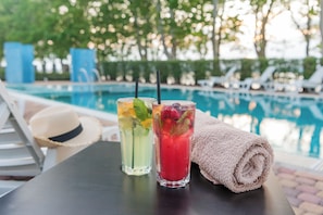 A poolside table with two drinks, a rolled towel, and a hat.