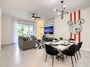 A modern living room with a sofa, a dining table set for four, a TV mounted on the wall, and a ceiling fan.