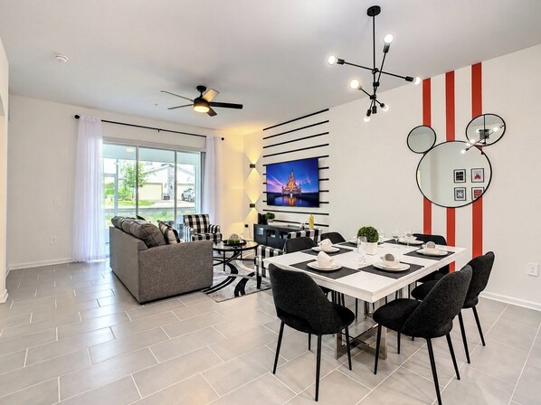 A modern living room with a sofa, a dining table set for four, a TV mounted on the wall, and a ceiling fan.