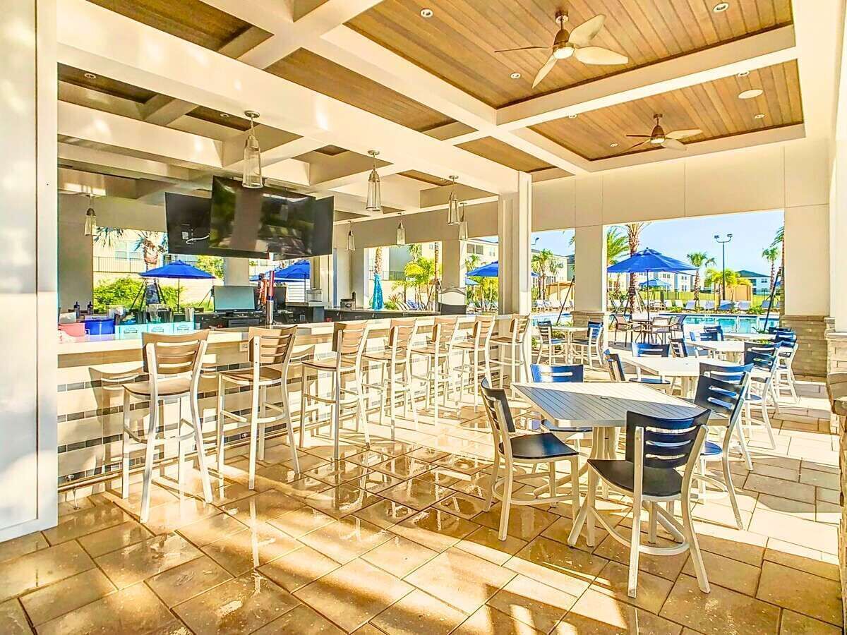 An outdoor dining area with multiple tables and chairs, a bar area with a TV, and a view of the pool and palm trees.