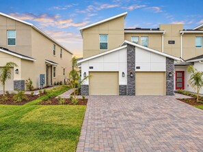 A modern residential building with a brick driveway, numbered garage doors, and a landscaped front yard.