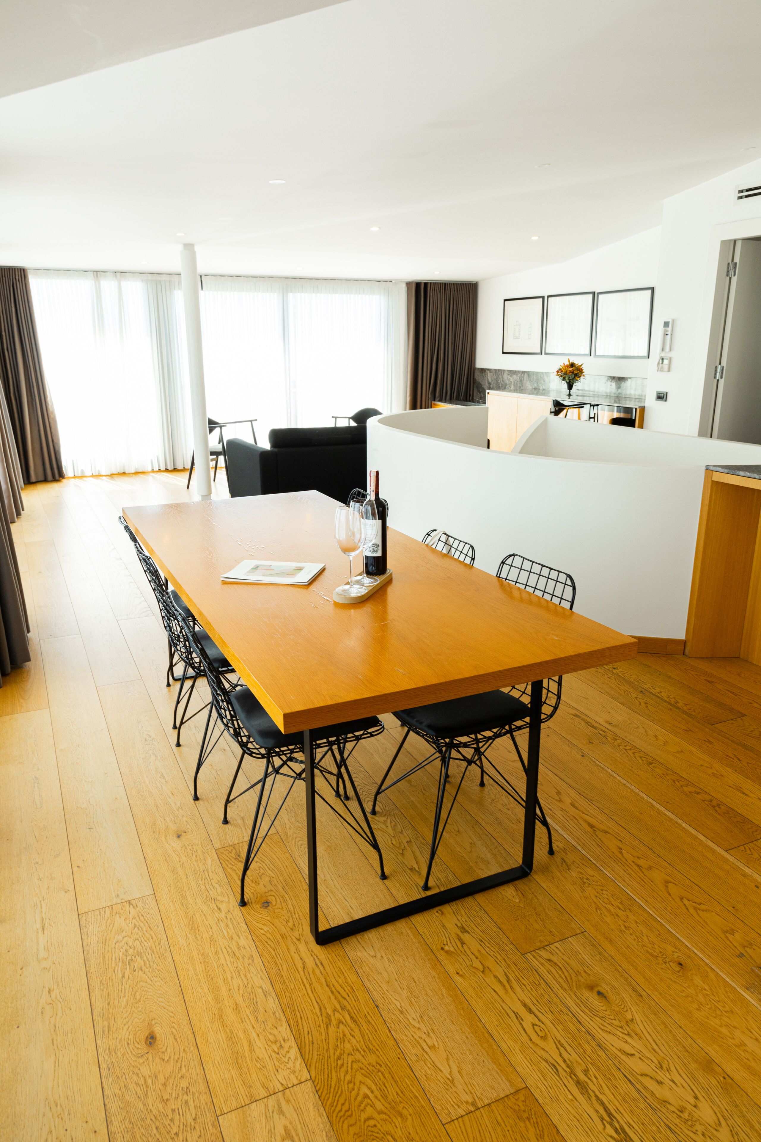 A modern hotel room with a wooden dining table, black chairs, a kitchen area, and large windows with curtains.