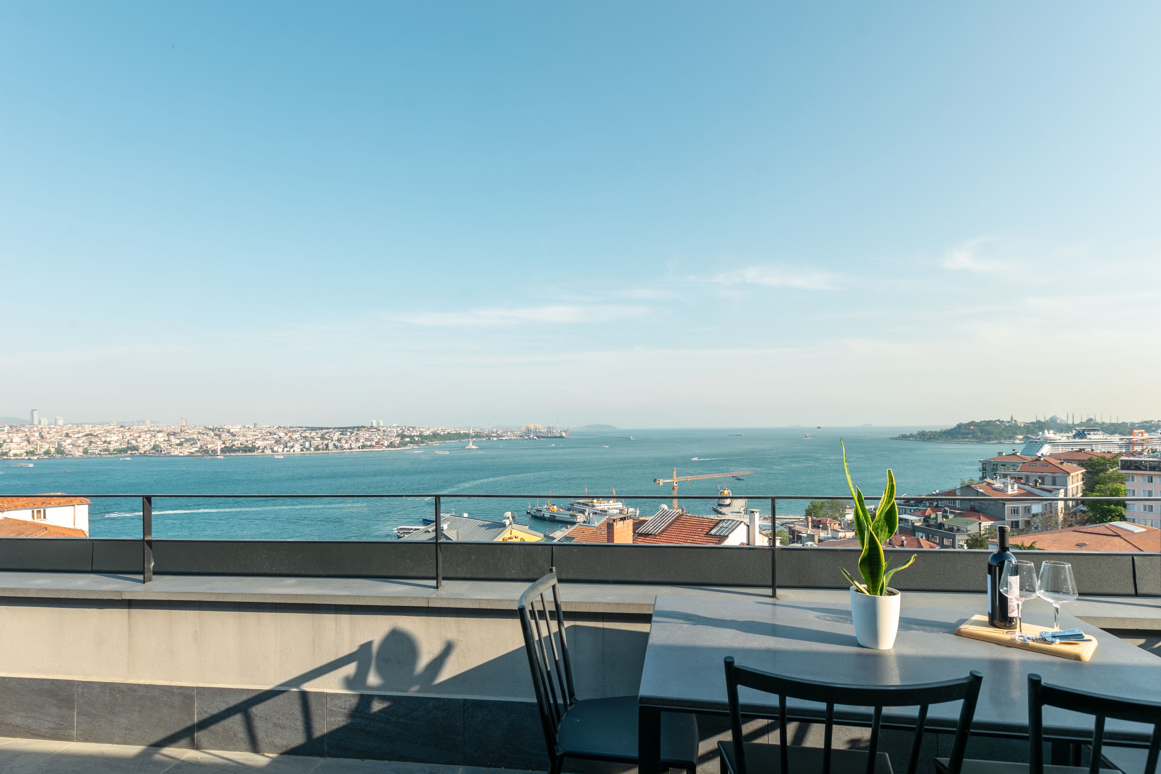 Een terras met een tafel, stoelen, een bloempot en uitzicht op zee en de stad.