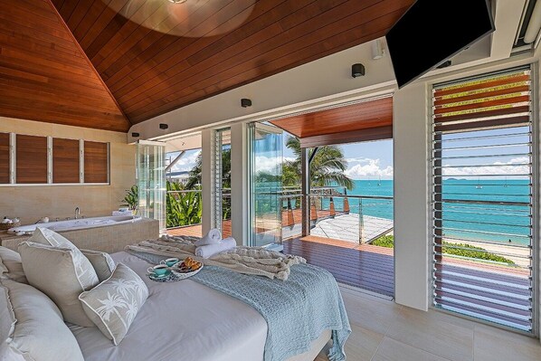 A bedroom with a large bed, a view of the ocean, and a balcony with a pool.