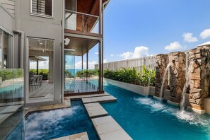 Un edificio moderno con zona de piscina cubierta de vidrio, terraza de madera y una cascada de piedra.