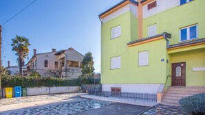 Un edificio residencial con fachada verde, una puerta principal y un pequeño jardín frontal con zona pavimentada y una cerca.