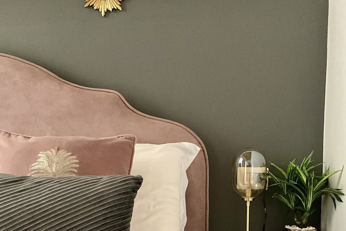 Un lit bien fait, avec une tête de lit rose, une lampe de chevet et une petite plante posée sur une table de chevet.