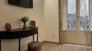 A room with a flat-screen TV mounted on the wall, a console table with decorative items, a potted plant, and a view through a door to an outdoor balcony. - Rua 39 fine apartments (Syracuse)