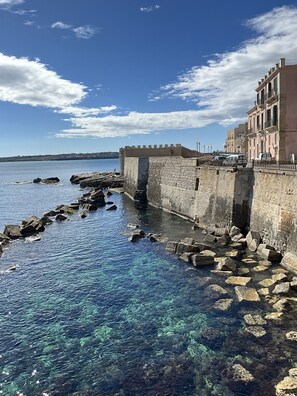 A coastal stone wall with clear water and a building with balconies. - Rua 39 fine apartments (Syracuse)