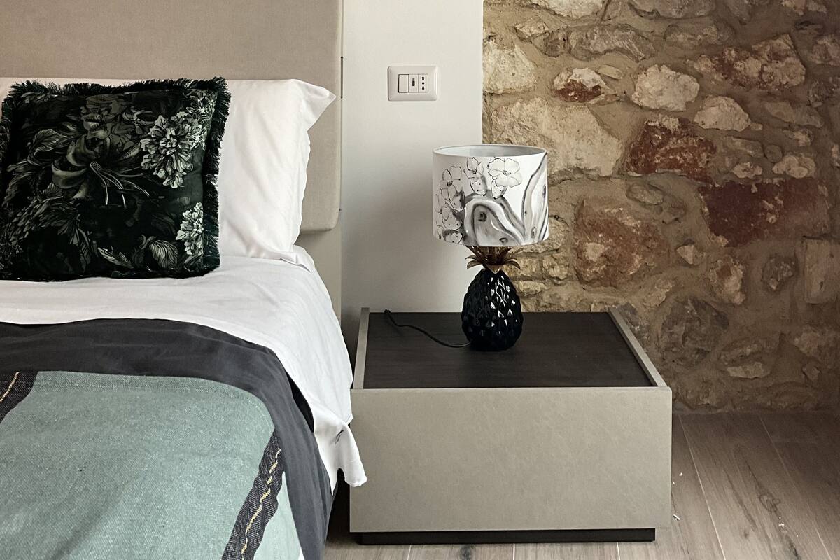 Une chambre à coucher comprenant un lit, une table de chevet, une lampe, une œuvre d’art murale et un mur en pierre.