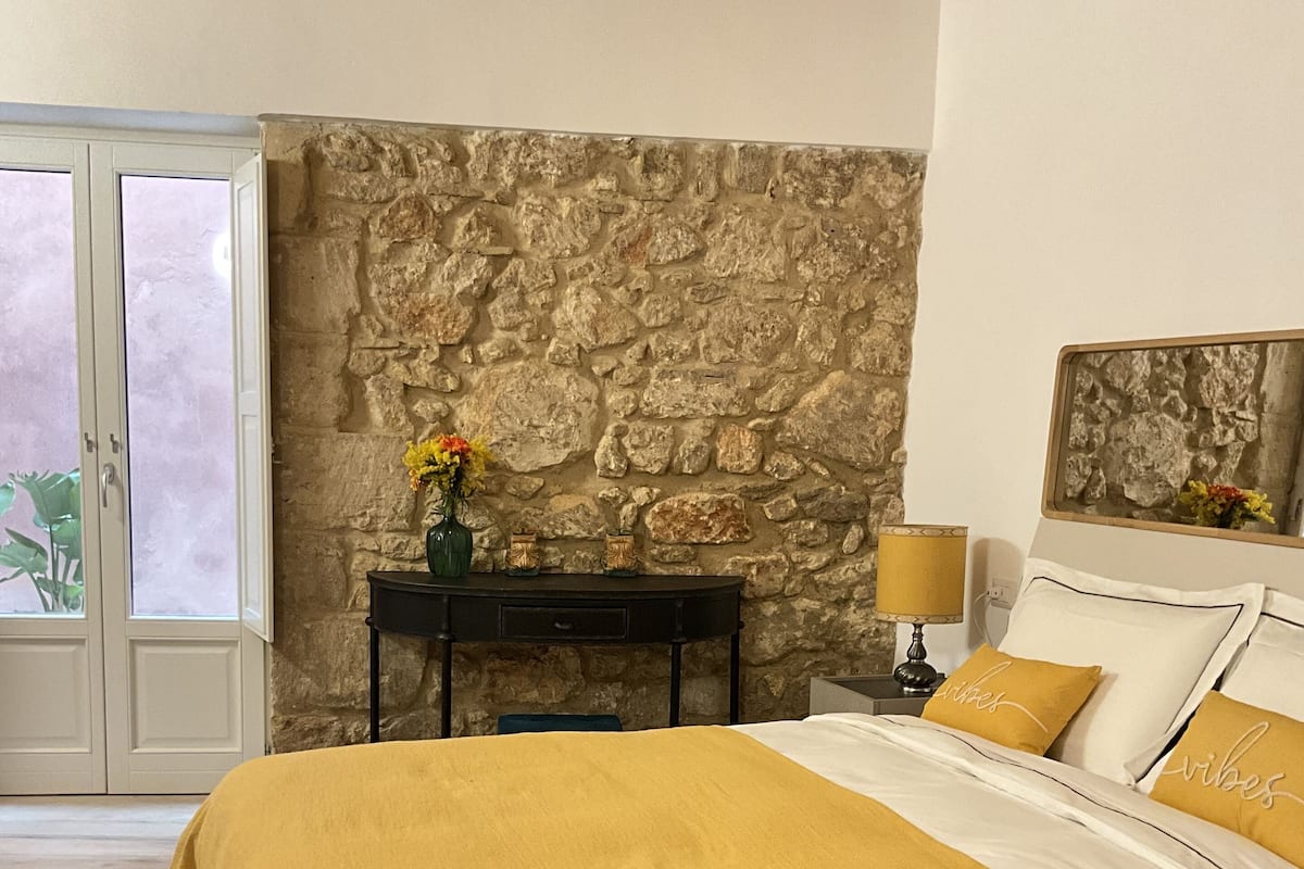 Une chambre avec un mur en pierre, un lit avec une literie jaune, une table de chevet avec une lampe et une fenêtre donnant sur un paysage verdoyant.