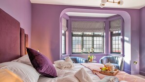 Ein Schlafzimmer mit einem großen Bett, violetten Wänden und einem Erkerfenster mit gemusterten Vorhängen.