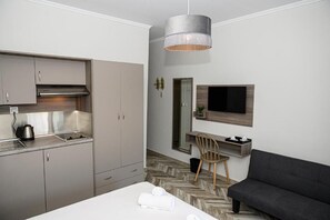 A modern hotel room with a kitchenette, a sofa, a TV mounted on the wall, and a herringbone-patterned floor.