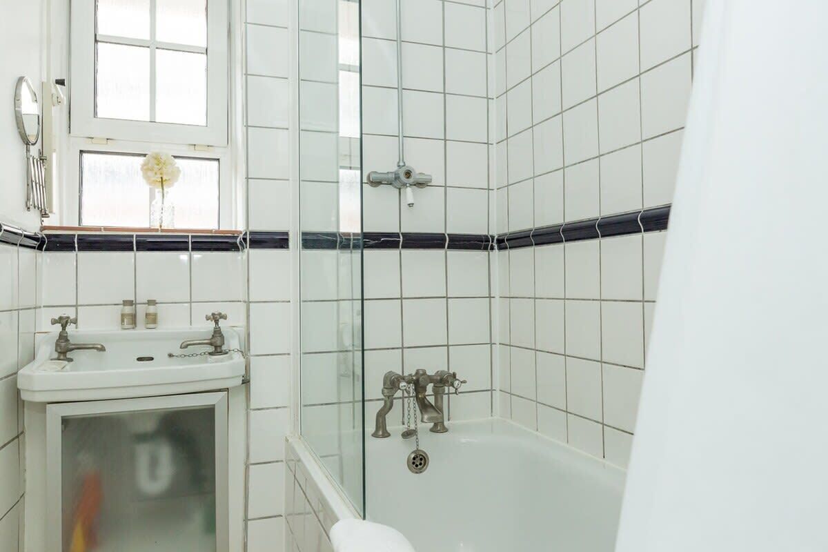Une salle de bain avec des carreaux blancs, une douche en verre, un lavabo blanc avec deux robinets et une fenêtre ornée d’une fleur.