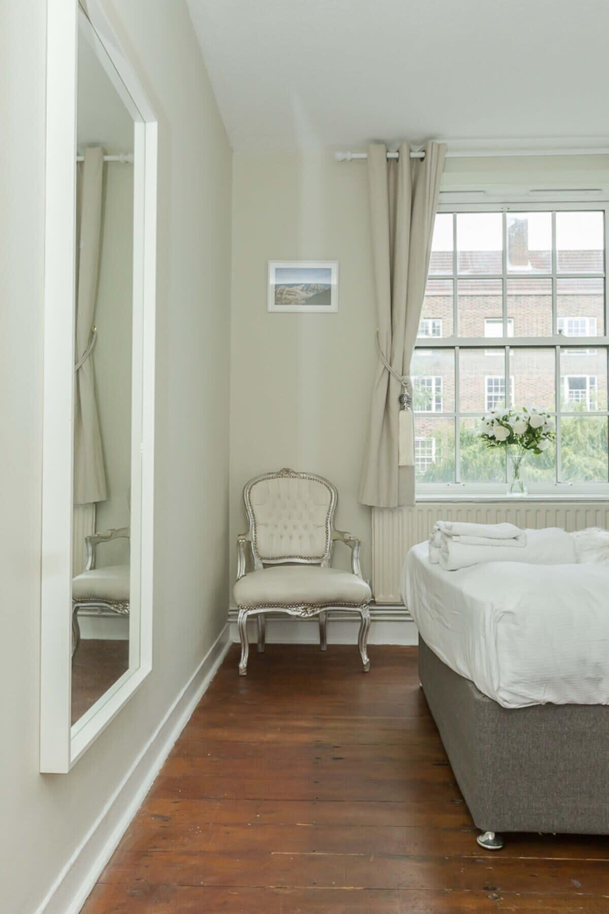 Une chambre à coucher avec un lit, une chaise, une fenêtre donnant sur des immeubles et un miroir.