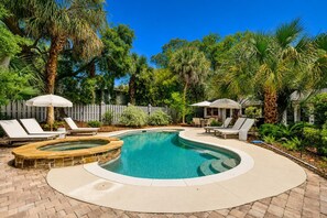 A swimming pool with a hot tub, surrounded by lounge chairs and umbrellas, with a wooden fence and lush greenery in the background.