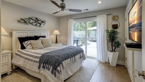 Un dormitorio con cama, mesitas de noche, ventilador de techo, ventana grande con cortinas, planta en maceta y televisor instalado en la pared.