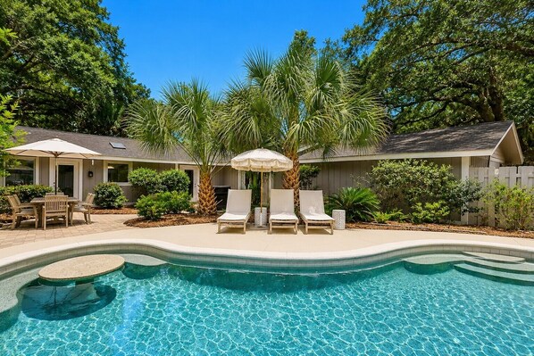 A swimming pool with lounge chairs and umbrellas, surrounded by a wooden fence and greenery.