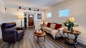 Un salon avec un canapé en cuir, une table basse en bois et un fauteuil à motifs. Une porte donne accès à une autre pièce, et il y a une fenêtre avec des stores.