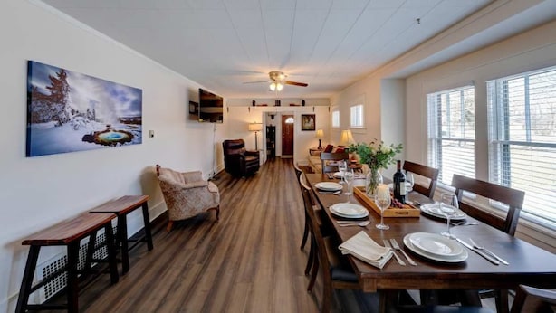 Un coin repas avec une table en bois dressée pour quatre personnes, un ventilateur de plafond et un tableau accroché au mur.