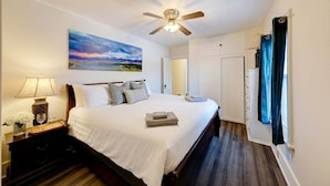 Une chambre à coucher avec un lit, un ventilateur de plafond, un tableau, une table de chevet, une lampe et une fenêtre avec des rideaux.