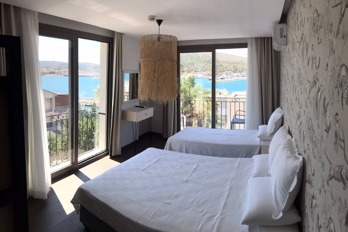 A hotel room with two beds, large windows with a view of a body of water and hills, a hanging light fixture, and a wall with a patterned wallpaper.