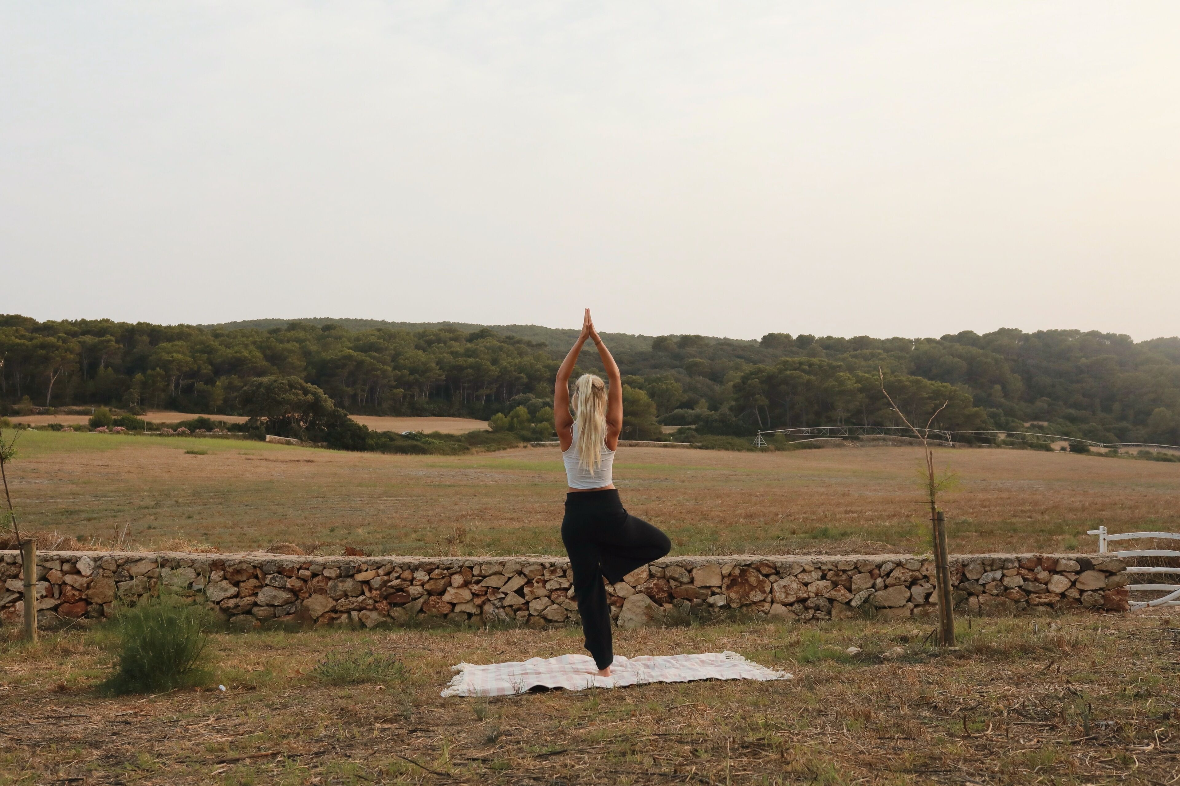 Una persona esegue una posa yoga su un tappetino in un campo aperto, con un muro di pietra e alberi sullo sfondo.