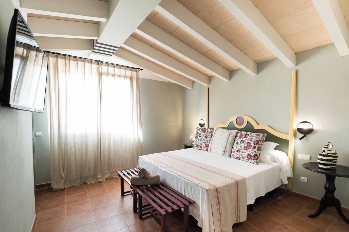 A bedroom with a bed, bench, bedside table, and a wall-mounted lamp.
