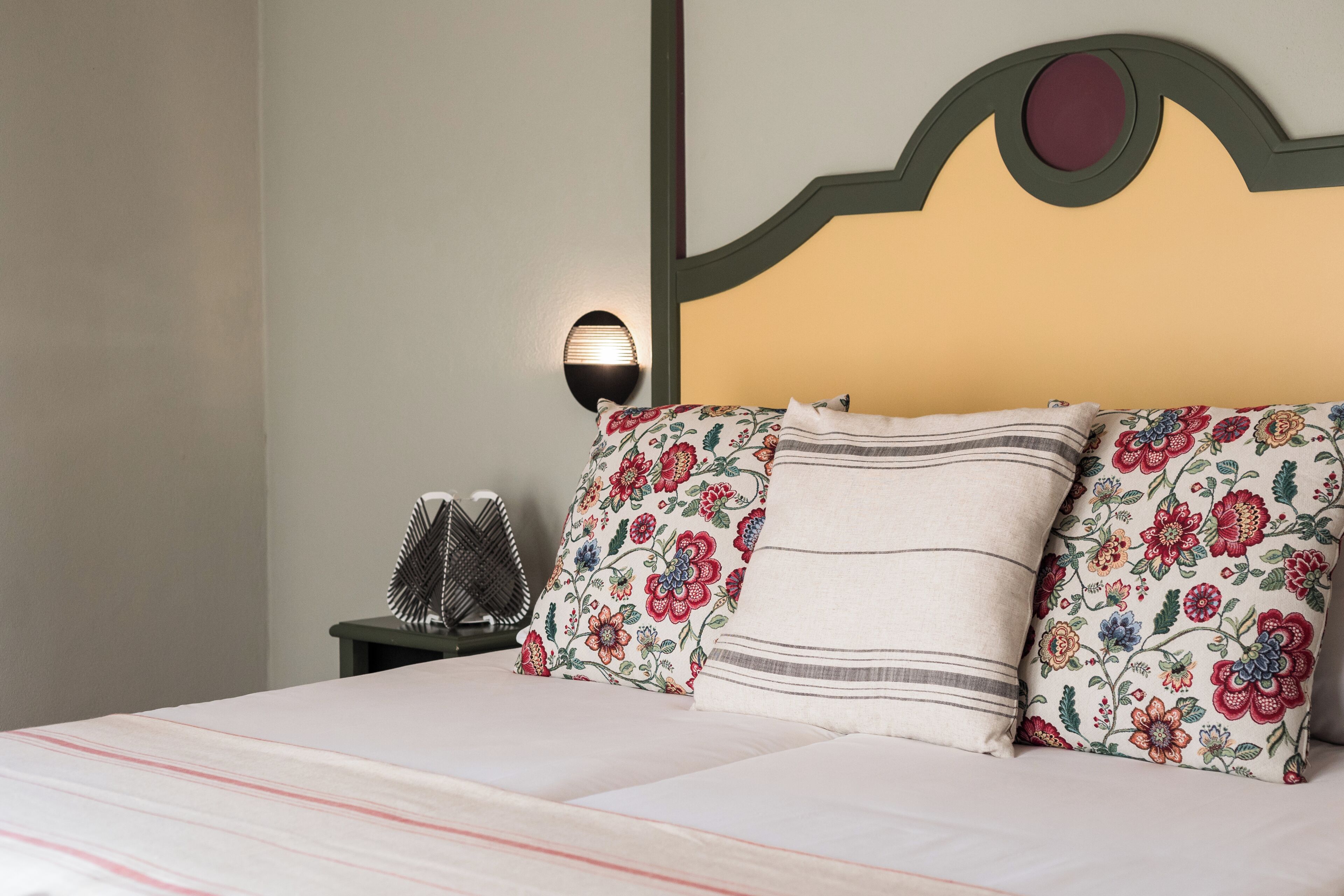 A bed with floral-patterned pillows, a headboard with a circular cut-out, and a wall-mounted lamp.
