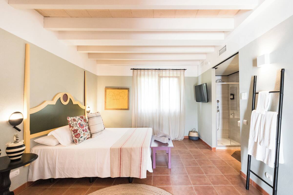 A hotel room with a bed, a small table, a chair, a shower area, and a television.