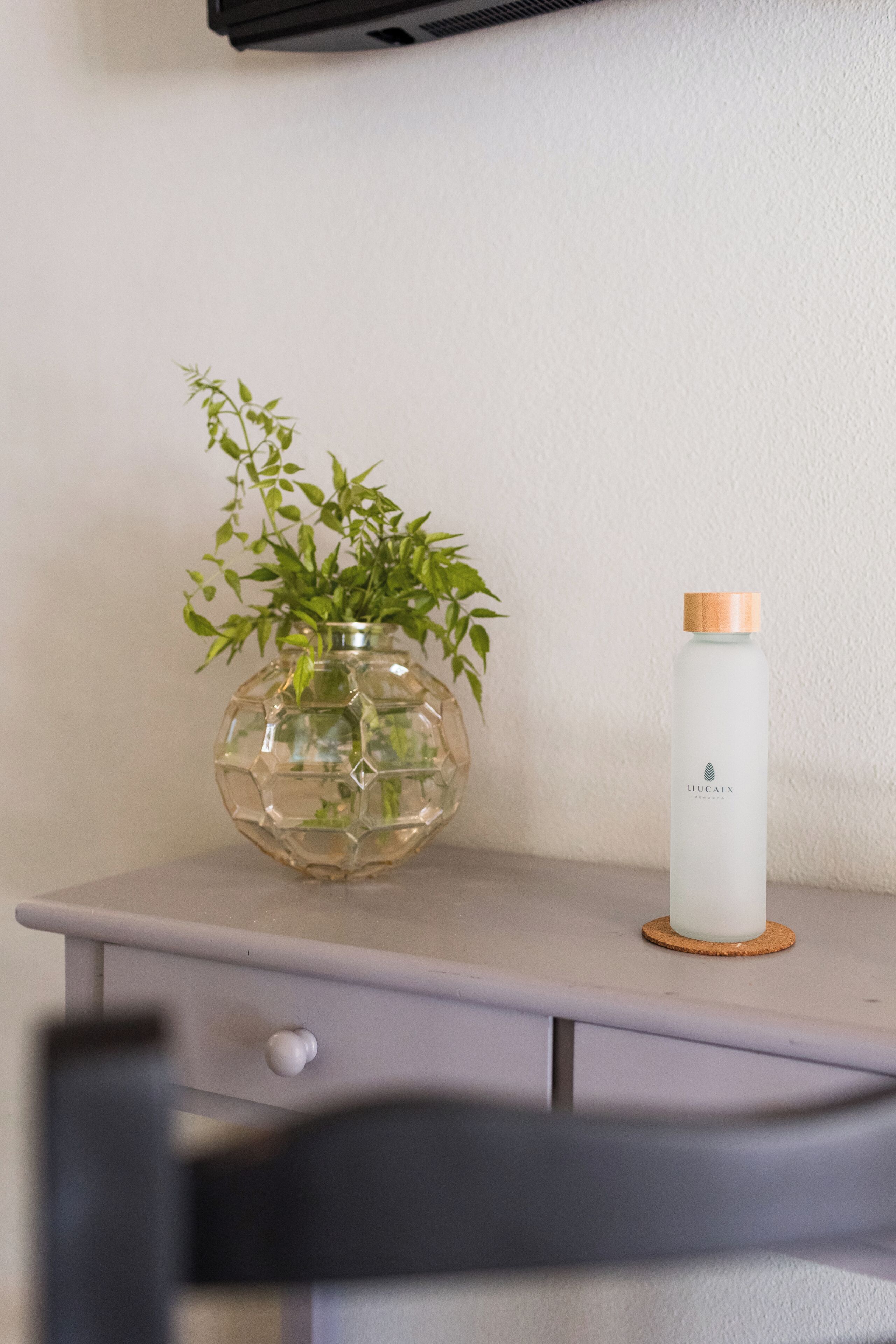A clear glass vase with green plants on a white wall, a white bottle with a wooden cap, and a grey drawer with a round knob.