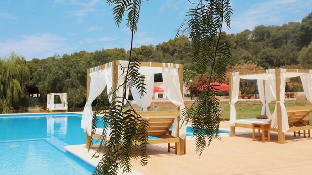 Uma área à beira da piscina com espreguiçadeiras de madeira e toldos, rodeada por vegetação e um céu claro.