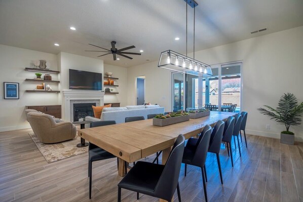 A modern living room with a fireplace, a dining area with a long wooden table and chairs, a ceiling fan, and a large TV mounted on the wall. - Arcadia 66 | PRIVATE POOL & HOT TUB! Arcade * Pool Table (Santa Clara)