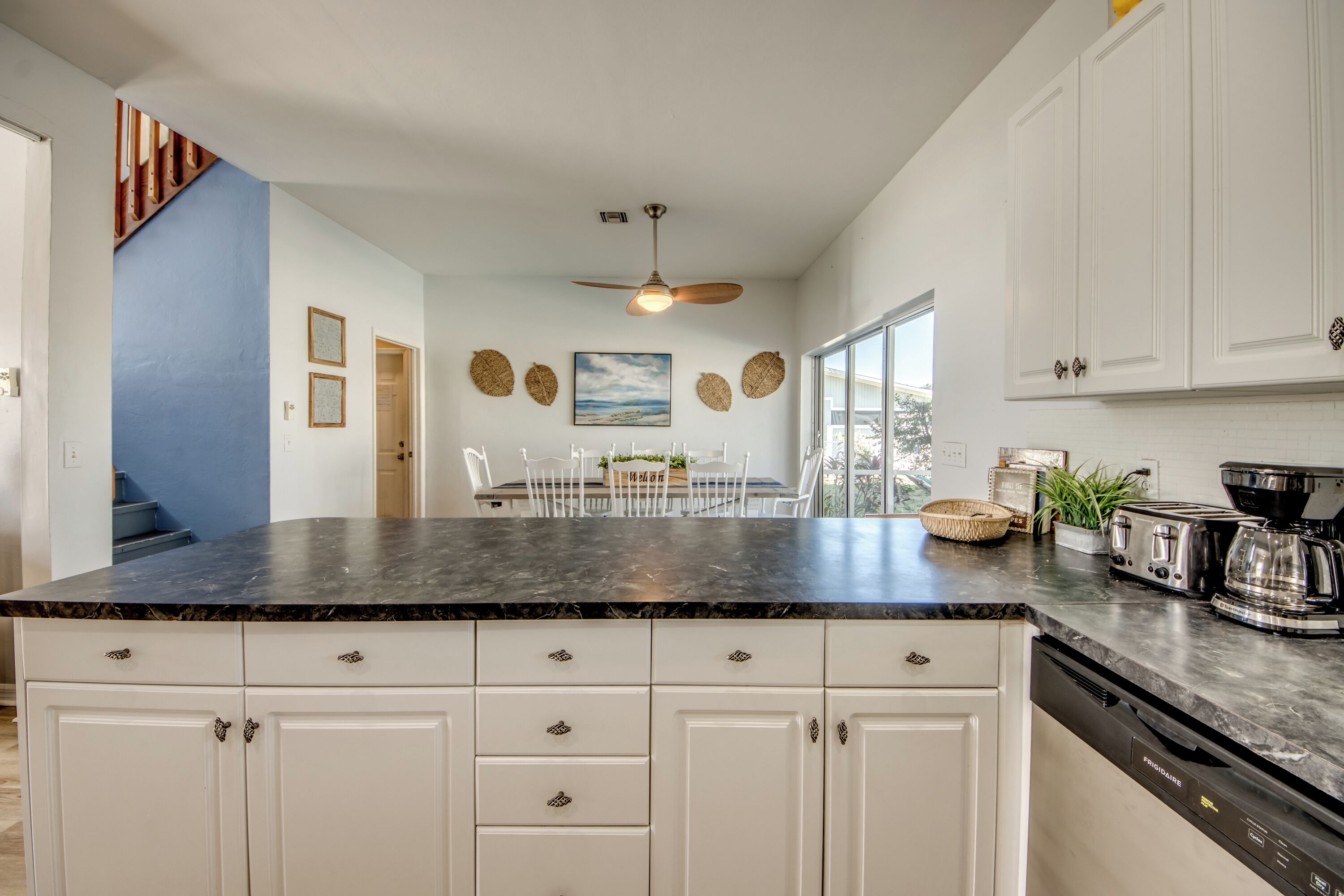 A modern kitchen with white cabinetry, a black marble countertop, and a ceiling fan. There is a dining area with a table and chairs, and a window providing natural light.