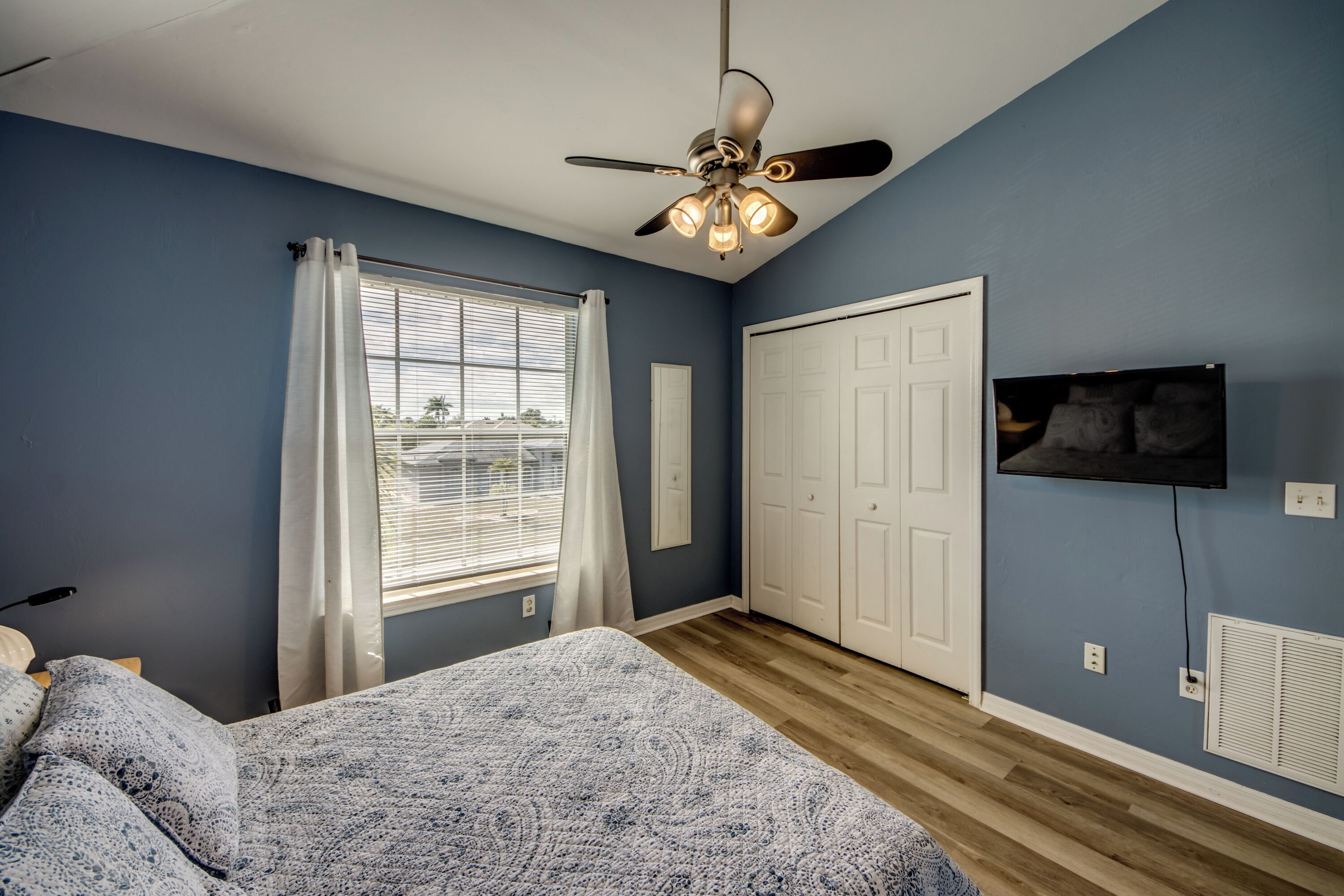 A bedroom with a bed, a ceiling fan, a window with curtains, a white door, and a wall-mounted TV.
