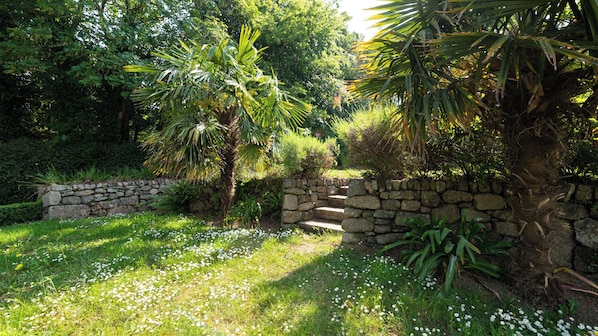 Una escalera de piedra que sube hacia un muro de piedra, rodeada de abundante vegetación y flores blancas.