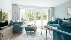 Una sala de estar moderna con sofá, sillón y mesa de centro, amplias puertas correderas de vidrio a un jardín y un televisor sobre un mueble de madera.