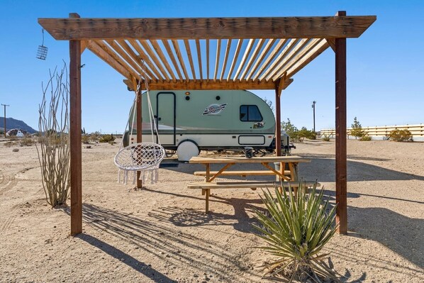 Ein Wohnmobil steht unter einer hölzernen Pergola mit einem Picknicktisch und einem Hängesessel.