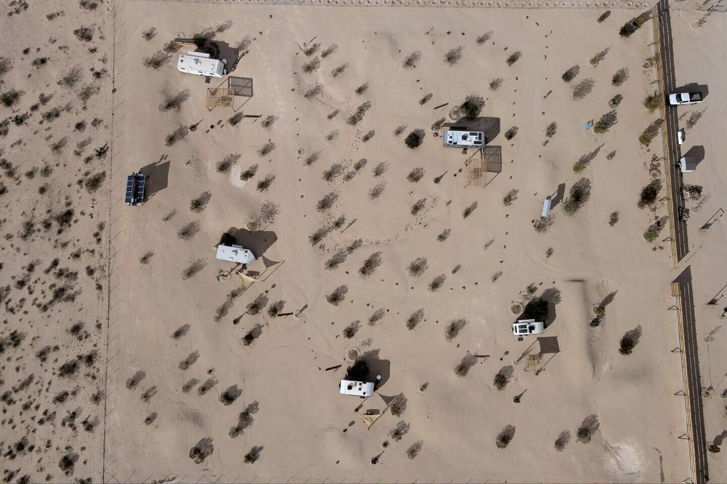 An aerial view of a desert area with several small buildings, some with attached vehicles, and a road running parallel to the buildings.
