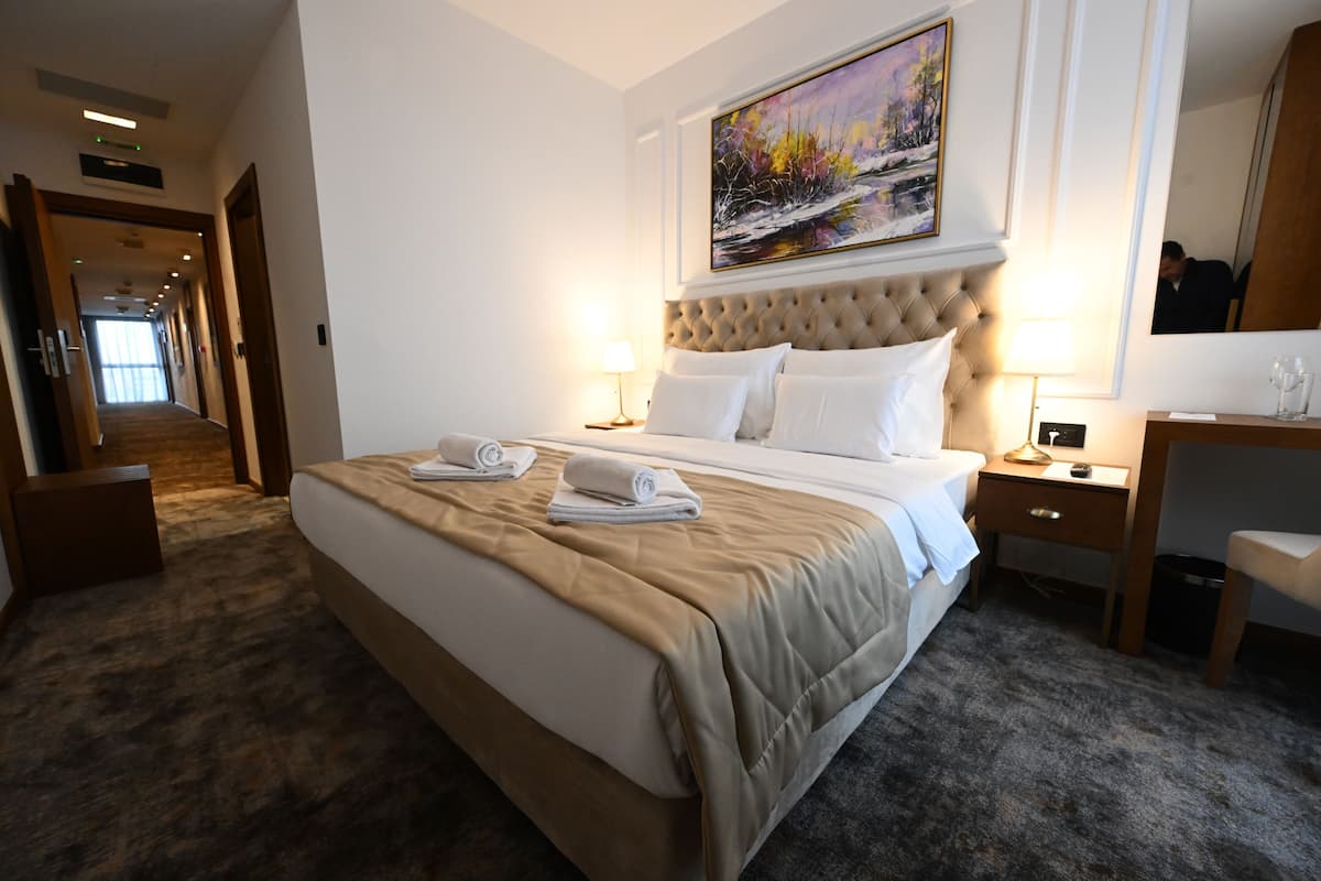 Une chambre d’hôtel avec un grand lit, deux tables de chevet, un bureau, une chaise, un tableau au mur et un couloir menant à d’autres chambres.