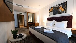 Habitación de hotel con una cama grande, un escritorio, una silla, una mesita con una planta y mandos a distancia, un espejo y un cuadro en la pared.