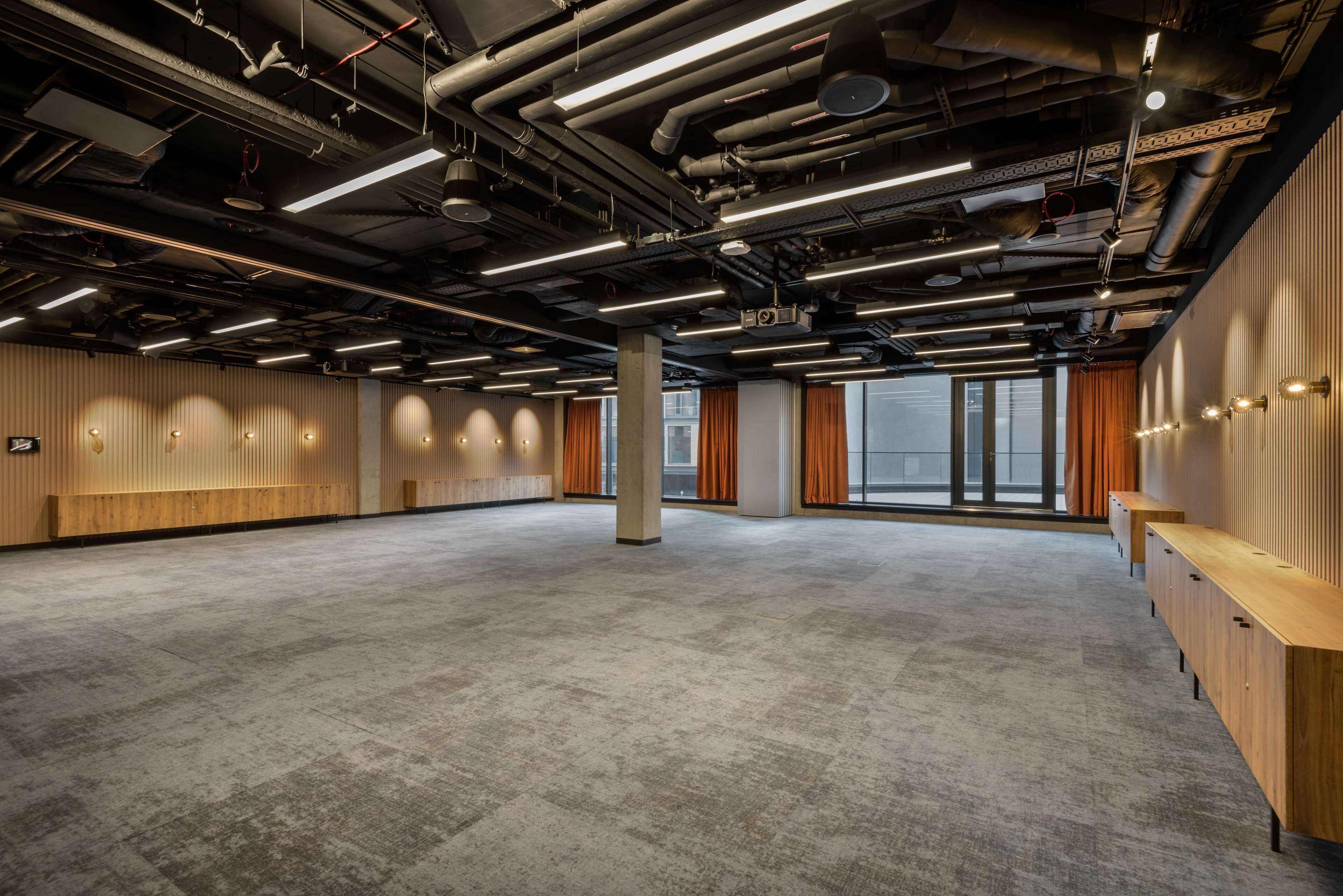Ein geräumiger, moderner Konferenzraum mit hoher Decke, freiliegenden Lüftungsrohren und einem großen Fenster, das natürliches Licht hereinlässt.