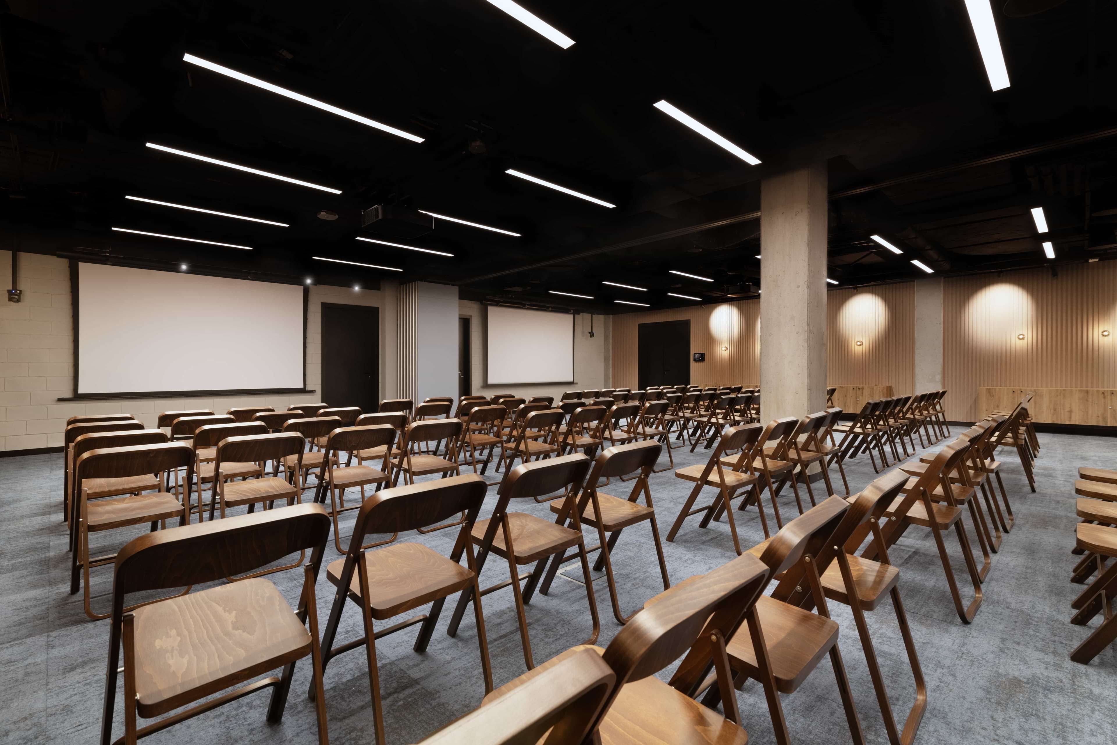 Konferenssitila, jossa on riveissä puutuoleja ja -pöytiä, projektorikangas ja modernit valaisimet.